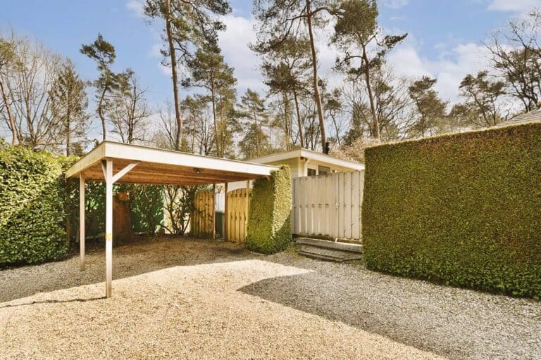 Wiaty garażowe drewniane — wolnostojący carport o płaskim dachu, na drewnianych słupach, na żwirowym podjeździe przy domu